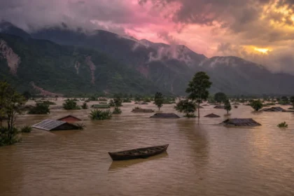 Submerged village in Northeast India during heavy monsoon floods, with rooftops above floodwater and hills behind